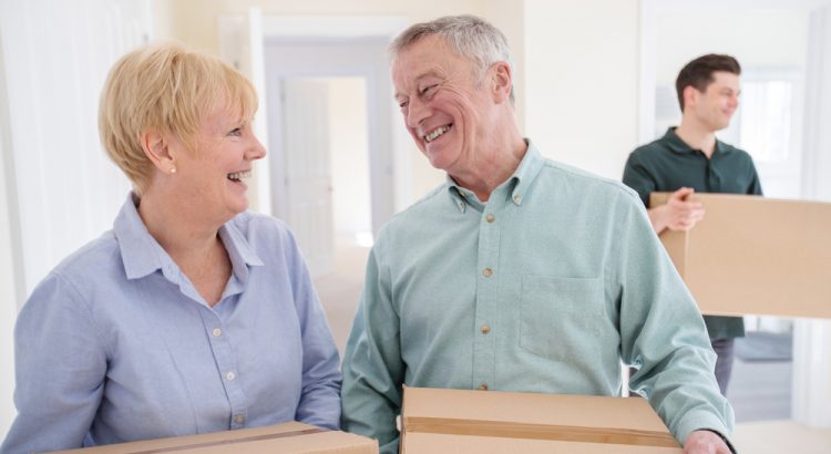 Senior Couple Downsizing In Retirement Carrying Boxes Into New Home On Moving Day With Removal Man Helping